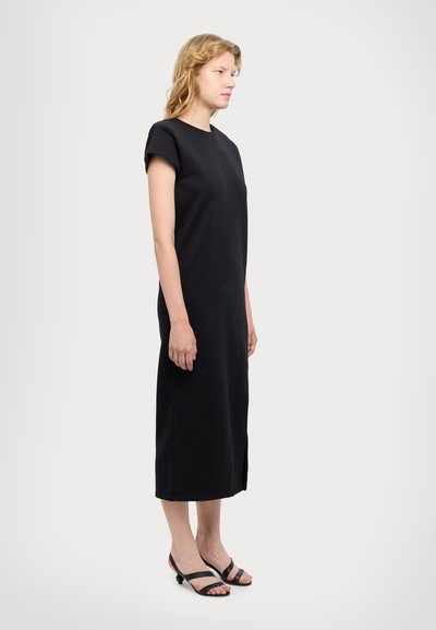 Robe noire, hauteur genoux, avec un col rond et des manches courtes, fabriquée en tissu doux. Détail de fente sur le côté, associée à des sandales à lanières noires.