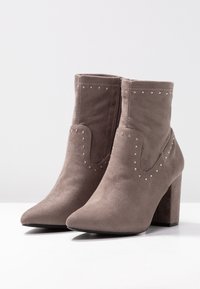 Botas de tobillo de ante gris con punta afilada y tacón ancho. Presentan tachuelas metálicas a lo largo de los laterales y un detalle de costura en la parte superior.