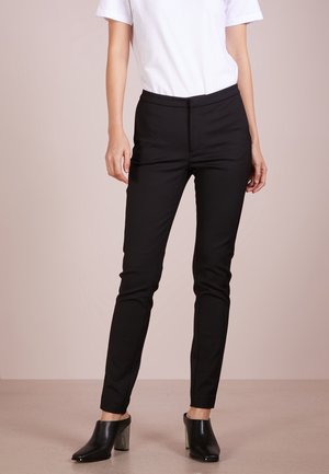 Person trägt schwarze Slim-Fit-Hosen, weißes kurzärmeliges Oberteil und schwarze Slip-on Pumps und steht vor einem einfarbigen Hintergrund.