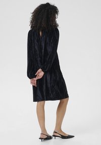 Rochie din catifea neagră cu mâneci lungi și detaliu cu cheiță la spate. Textură netedă și croială lejeră, asortată cu pantofi slingback negri.