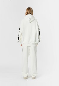 Sudadera blanca con capucha y acentos gráficos negros en las mangas, combinada con pantalones blancos sueltos. El material parece suave y texturizado.