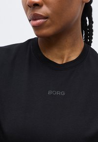 Κοντινό πλάνο ενός ατόμου που φοράει μαύρο μπλουζάκι με στρογγυλή λαιμόκοψη και τη λέξη "BORG" τυπωμένη με μικρά, σκοτεινά γράμματα στο στήθος, πλεξούδες μαλλιών ορατές στο πλάι.