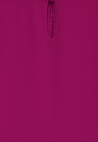 Maglietta fucsia con tessuto testurizzato, dotata di un piccolo foro a chiave sulla parte anteriore chiuso da un bottone. Finitura liscia complessivamente.