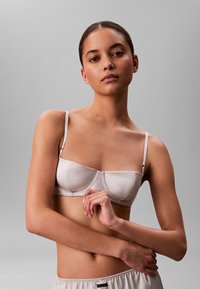 Jasnoróżowy biustonosz o usztywnionej konstrukcji, cienkich regulowanych ramiączkach i gładkiej satynowej fakturze. Wyeksponowany na szarym tle.