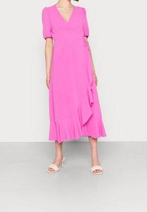 Różowa sukienka o kroju wrap z krótkimi bufkami, dekoltem w kształcie litery V i falbanym dołem. Gładki materiał oraz wiązanie w talii. Noszona z kremowymi sandałami na słupku.