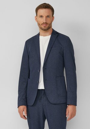 Man met baard, gekleed in een marineblauw colbert, bijpassende broek en een wit T-shirt, staand tegen een effen lichtgrijze achtergrond.