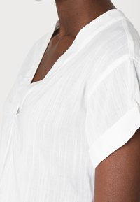 Blusa bianca in lino con scollatura a V, maniche corte arrotolate e sottili righe verticali. Tessuto leggero con una vestibilità rilassata.