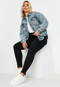 Jeansjacka med ljus tvätt, knäppt fram, och två bröstfickor, i kombination med svarta tighta byxor och vita sneakers, sittande på en plattform.