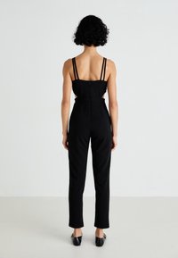 Woman with short curly hair wearing a sleeveless black jumpsuit with cutouts and black slip-on shoes, standing against a white background.