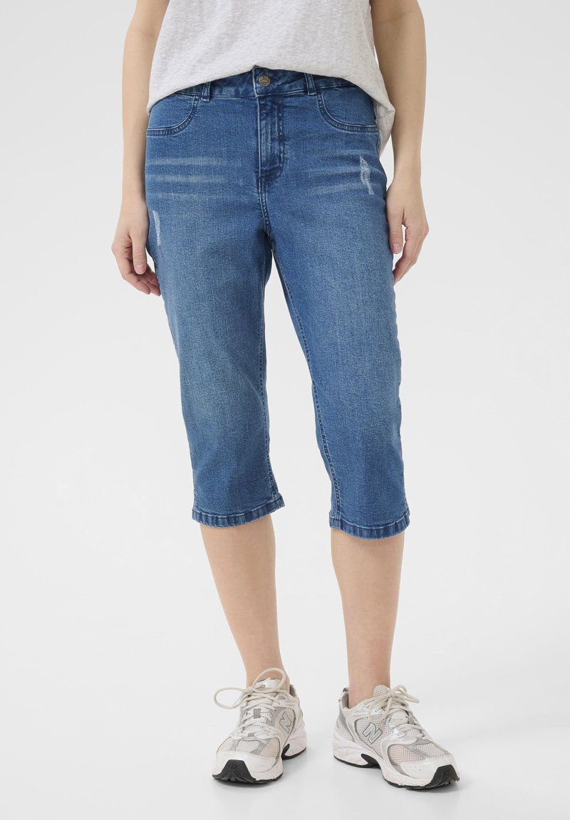 Jeans capri-byxor i medelblått med rak skärning, knälång längd och subtila slitningar. Styld med en grå topp och sneakers.