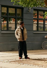 Doudoune beige avec capuche, deux poches latérales, portée sur un chemisier zippé noir, associée à un pantalon noir ample et des chaussures marron.