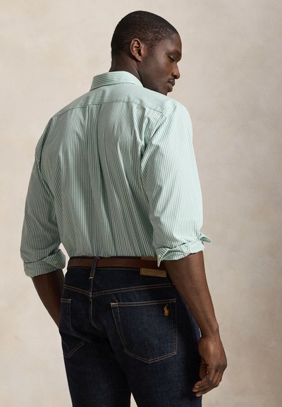 Mężczyzna w jasnografitowej koszuli w zielone paski z długim rękawem podwiniętym, ciemnoniebieskich dżinsach z brązowym paskiem, stoi tyłem i patrzy w prawo.