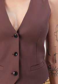 Gilet marron avec un profond col en V, doté de quatre boutons noirs et de deux poches latérales, fabriqué en tissu lisse avec peu de détails de texture.