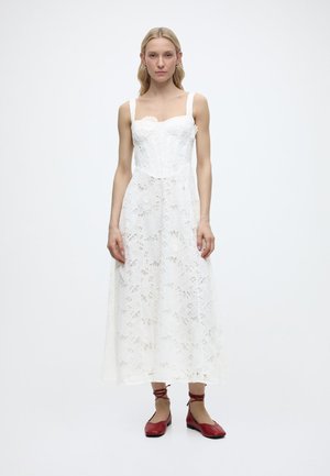 Γυναίκα που φοράει λευκό φορεμα με δαντέλα και λουλούδια, χωρίς μανίκια, μήκους μέχρι τη μέση της γάμπας, με τετράγωνο ντεκολτέ και κόκκινα επίπεδα παπούτσια με κορδόνια γύρω από τον αστράγαλο, στέκεται σε απλό λευκό φόντο.