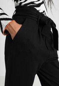 Zwarte, hoge taillebroek met een stoffen riem en strikdetail, voorzien van zijzakken en een gladde textuur. Zichtbare tailleband en slank ontwerp.