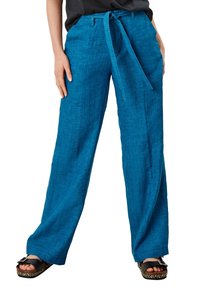 Pantalon large en lin turquoise avec ceinture à nouer, surface texturée, poches latérales et coupe décontractée. Sandales noires portées.