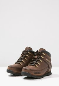 Brązowe skórzane buty do wędrówek z czarnymi akcentami, textured surface, metalowe oczka i gumowa podeszwa. Zawierają branding po bokach.