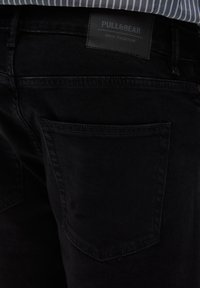 Jean noir en denim au design épuré, avec une poche arrière unique et une étiquette marquée "PULL&BEAR DENIM COLLECTION" au niveau de la taille.