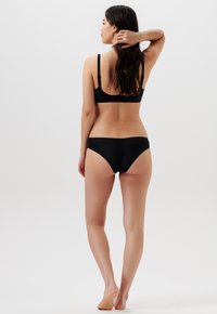 Vrouw die blootvoets staat, met haar rug naar de camera gekeerd, gekleed in een zwarte beha en doorschijnend gestreept zwart ondergoed, met één arm opgeheven om haar haar aan te raken.