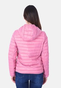 Donna con lunghi capelli scuri che indossa una giacca a cappuccio imbottita rosa e jeans blu, vista di schiena su uno sfondo bianco.