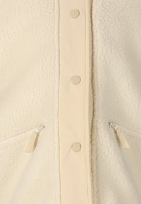 Cremefarbener Fleece-Stoff mit zentralen Druckknöpfen und zwei diagonalen Reißverschlusstaschen.
