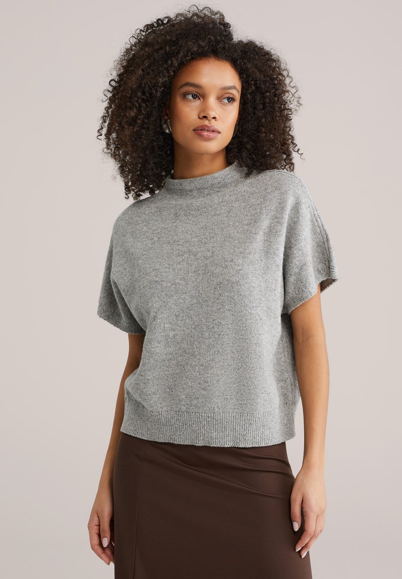 Pull gris à manches courtes en tricot avec un col montant et un ourlet côtelé, présentant une texture douce et une coupe décontractée, associé à une jupe marron.