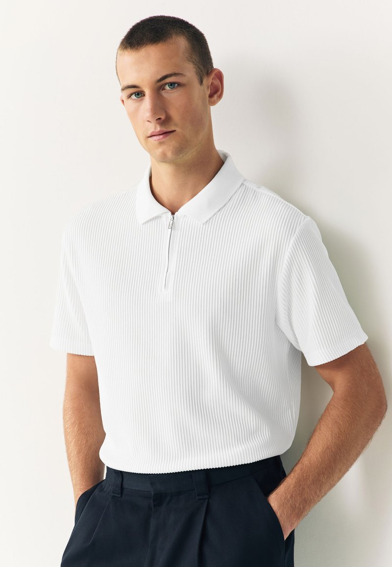 Young man with short hair wearing a white ribbed zip-up polo shirt and black pants standing against a white wall.