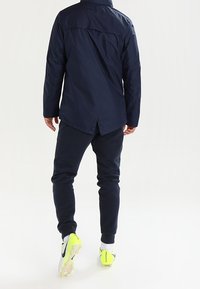 Giacca lunga a maniche lunghe di colore blu navy, leggera e con una vestibilità dritta. Abbinata a pantaloni jogger scuri e scarpe da corsa gialle con dettagli neri.