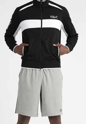 Mężczyzna w czarno-białej kurtce zapinanej na zamek z logo Polo i jasnoszortach z logo Polo, stojący z rękami w kieszeniach kurtki.