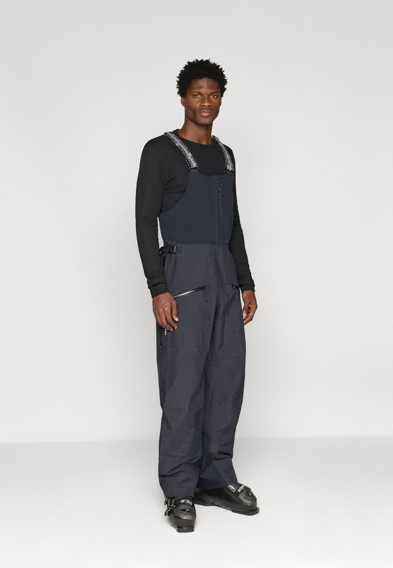 Sötétkék anyagból készült síelős overall, állítható pántokkal, cipzáras elejével és több cipzáras zsebbel a praktikum érdekében.