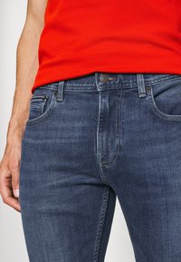 Vaqueros de mezclilla azul oscuro con un corte regular, que presentan una cremallera, diseño de cinco bolsillos y detalles de costura sutiles.