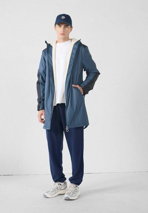 Giacca impermeabile blu con fodera crema e dettagli neri, abbinata a pantaloni jogger blu navy e sneakers grigie; presenta una cerniera frontale e cappuccio.