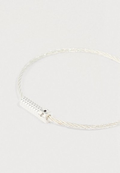 Strieborný zapletaný drôtový náramok s textúrovaným valcovým zapínaním označeným "11g" na jednofarebnom svetlom pozadí.