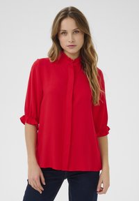 Blusa rossa con bottoni, colletto a volant e maniche a tre quarti. Tessuto morbido con una consistenza liscia e vestibilità comoda.