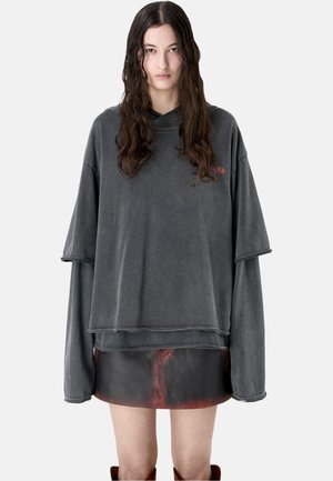 Młoda kobieta z długimi ciemnymi włosami, ubrana w luźną, wyblakłą szarą bluzę z kapturem i krótką czarno-czerwoną skórzaną spódnicę na jednolitym tle.