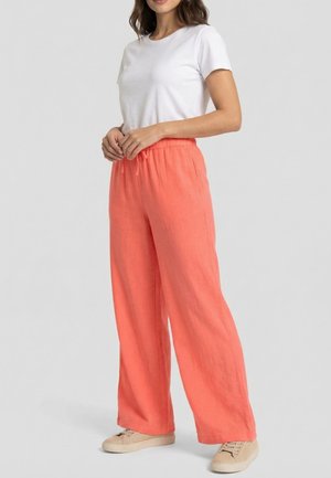 Vrouw draagt losse koraalkleurige broek met trekkoord in de taille, wit T-shirt met korte mouwen en beige sneakers, staand met handen geklemd.