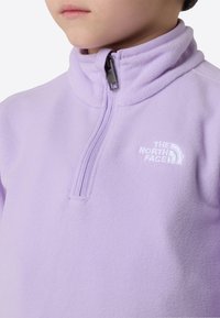 Lekki pulower z polaru w kolorze jasnej lawendy z przednim zamkiem na pół. Posiada wysoki kołnierz i wyszywane białe logo na lewej piersi. Miękka tekstura.