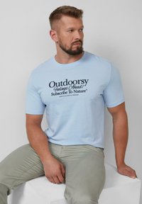 Helleblaue Baumwoll-T-Shirt mit kurzen Ärmeln und schwarzem Text: "Naturgefühle voraus! Abonnieren Sie Natur®" auf der Vorderseite.