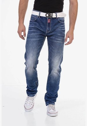 Uomo che indossa jeans blu sbiaditi con una cintura bianca con la scritta "CBJ" e sneaker bianche, in piedi davanti a uno sfondo bianco uniforme.