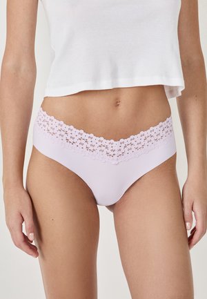 Femme portant un débardeur blanc court et un sous-vêtement léger violet avec une ceinture en dentelle florale, se tenant devant un fond neutre.