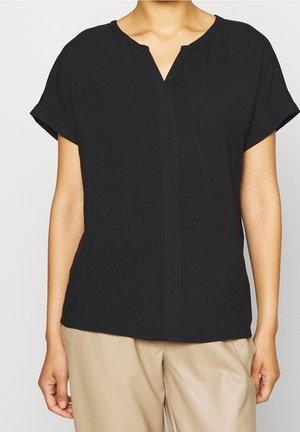 Femme portant un haut noir à manches courtes avec une petite encolure en V, associé à un pantalon beige, montrée des épaules aux hanches.