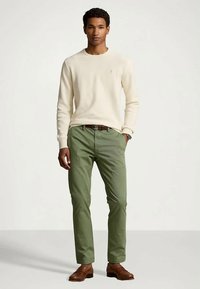 Mężczyzna stojący z rękami w kieszeniach, ubrany w kremowy sweter, oliwkowe spodnie, brązowy pasek, brązowe buty i zegarek na lewym nadgarstku.