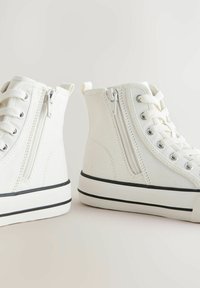 Witte canvas hoge sneakers hebben zijritsen, vetersluiting aan de voorkant, zwarte rubberen zolen en een gestructureerde afwerking.