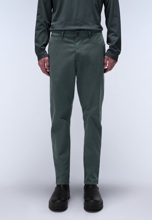 Man wearing dark green slim-fit pants, long-sleeve dark green shirt, and black slip-on shoes standing against light background.