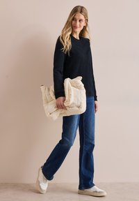 Marinefarbener strukturierter Pullover, leichte, gesteppten cremefarbene Tasche, blaue gerade Jeans und weiße Sneakers. Einfaches, lässiges Outfit vor neutralem Hintergrund.