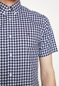 Hombre vestido con una camisa de botones de manga corta a cuadros en azul marino y blanco, con un pequeño logo en el pecho, frente a un fondo plano.