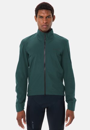 Man met een donkergroene jas met rits en zwarte fietsbroek, kijkt recht vooruit met een neutrale uitdrukking tegen een effen achtergrond.