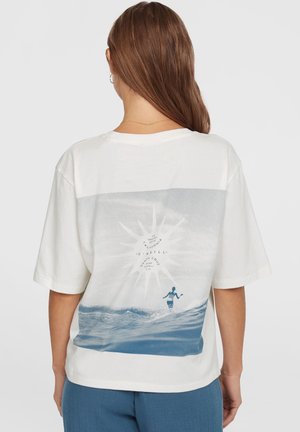 Kvinde iført hvid t-shirt med blå bølge og surfer grafik på ryggen, kombineret med blå bukser, vendt væk mod en ensfarvet baggrund.
