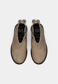 Une paire de bottines en suède beige avec des semelles noires, des zips sur les côtés et des tirettes décoratives, vues d'en haut sur un fond blanc.