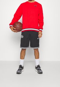 Červená mikina s dlouhým rukávem a logem, černé sportovní šortky s bílými detaily a textem, a šedé tenisky s černými prvky. Drží basketbal.